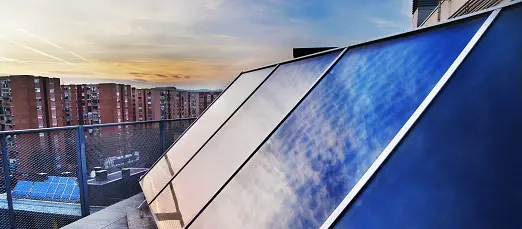 Solar Panels on a terrace of a building in a city at sunset. The panels reflect the blue sky. There are other buildings in the background.